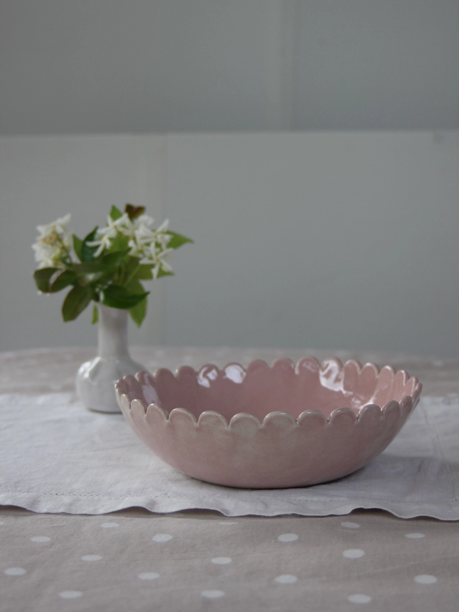 small pink serving bowl with scalloped edges and a vase of flowers on a tablescape made for serving vegetables or snacks, salads or fruit