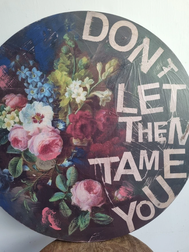 Art Panel made from a round table top. A dark background with multicolour flowers and writing in pink that says 'don't let them tame you'.