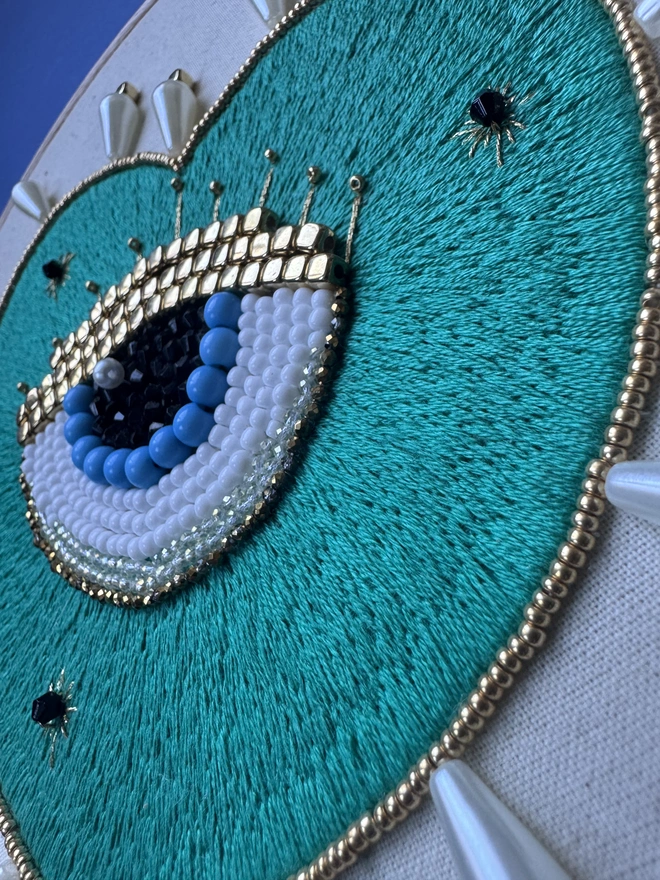 An embroidery hoop containing an embroidered hoop with a beaded eye and framed in gold and pearl beads