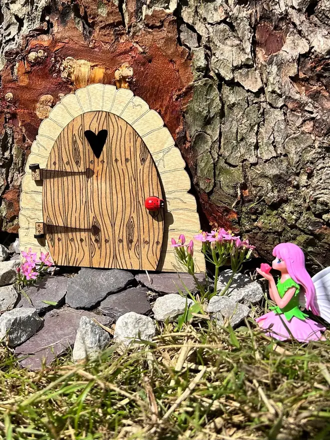 Wooden fairy door for kids with engraved woodgrain detail, heart-shaped window, and decorative hinges, displayed at the base of a tree surrounded by moss and wildflowers.