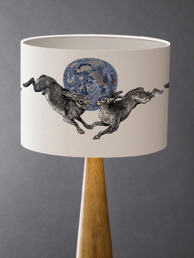Drum Lampshade featuring a pair of hares leaping across a blue and silver moon with a white inner on a wooden base 