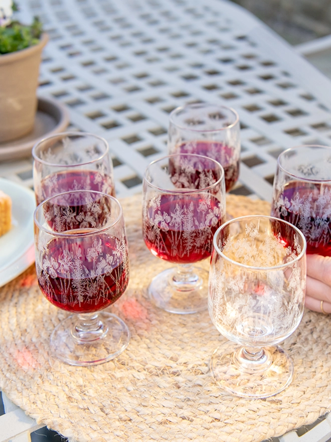 A short-stemmed bistro glass decorated with, illustrated poppies, hydrangeas, and sunflowers against a garden background.