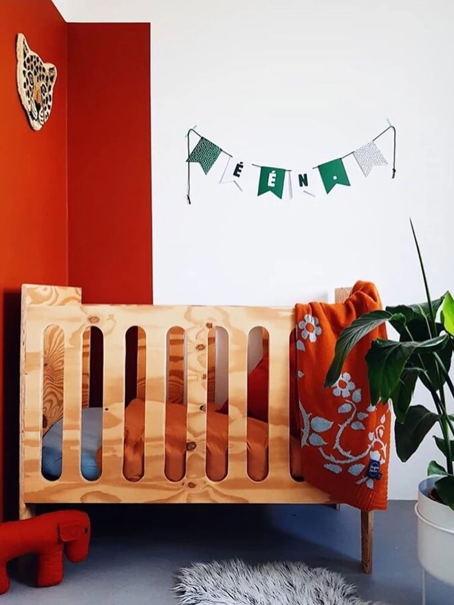 A modern nursery showing a wooden cot with a rust briar rose baby blanket thrown over the side. The walls are half painted in the same colour orange and a tiger head on the wall and large plant give jungle vibes.