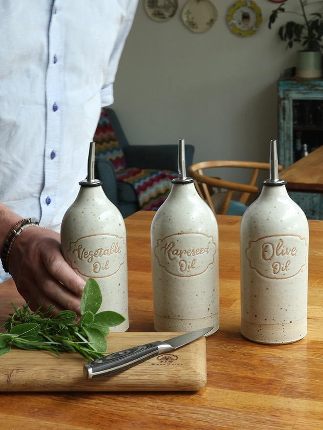 Three handmade stoneware oil pourers sat on a wooden table in a kitchen scene. The oil pourers feature the  words 'Vegetable Oil', 'Rapeseed Oil' and 'Olive Oil' and are glazed in an off-white glaze. A man is picking up the oil pourer labeled 'Vegetable Oil'