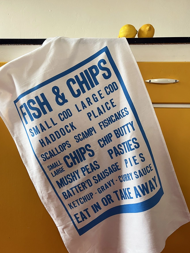Mr.PS Fish & Chips menu screenprinted tea towel hanging on a yellow cabinet