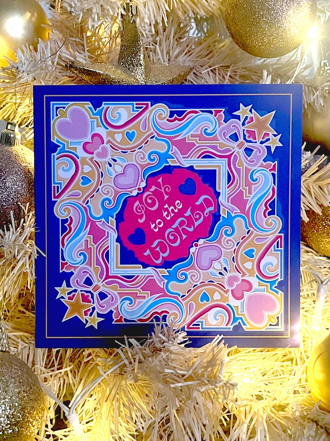 A square, blue Christmas card with an abstract, multi-coloured design including stars and hearts, with “Joy to the World” at the centre, rests in white and gold tinsel and baubles.