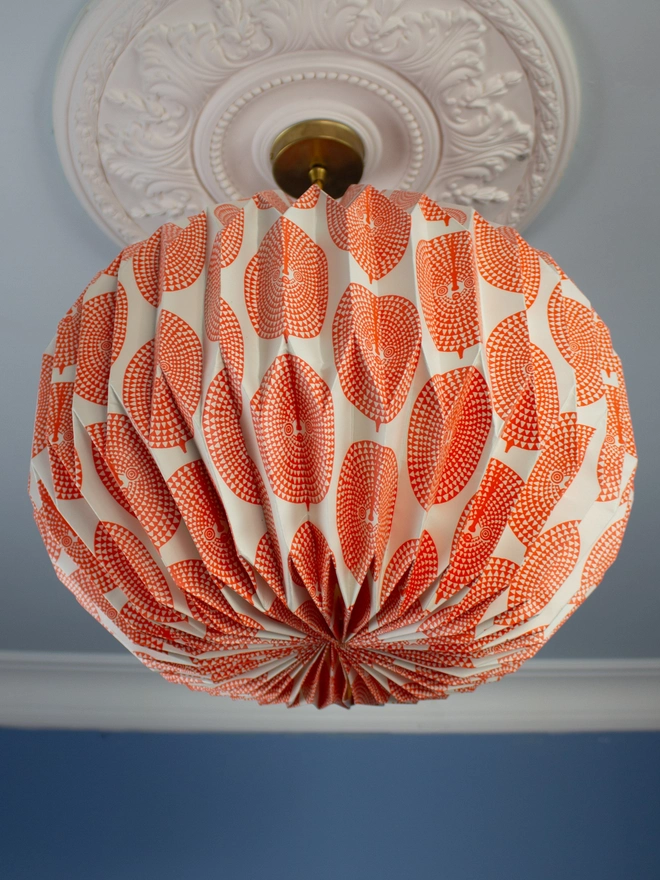 A vintage-inspired paper lampshade with a bold geometric pattern adds a touch of retro flair to a room with a blue wall and ornate ceiling. The lampshade is made from folded paper, creating a unique and eye-catching design.