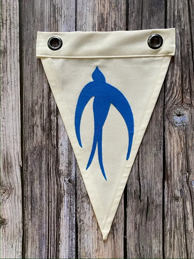 an ivory pennant flag with a blue swallow stitched on,  laying on a wooden floor with 