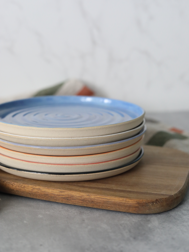 A stack of five handmade ceramic side plates on a wooden board, topped with a blue plate showing a concentric swirl texture, with coloured rim edges visible beneath in lavender, mandarin orange, cherry red, and black, styled with a linen cloth against a marble background