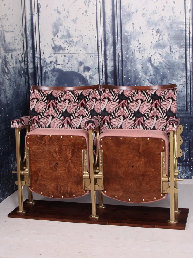 Set of two vintage cinema seats upholstered in a pink ostrich Art Deco design velvet with pink panel and piping against a blue marbled wall.  Seats are both flipped up