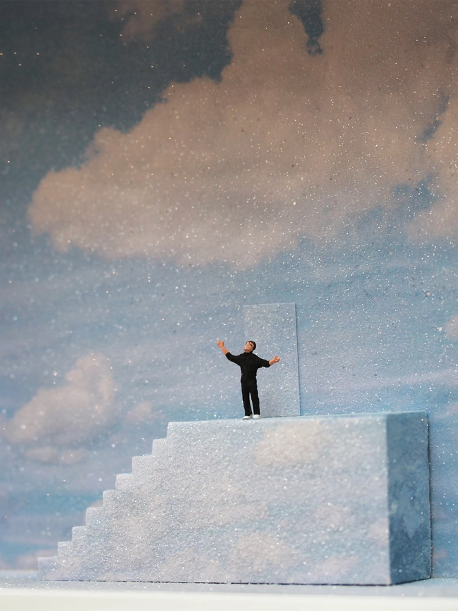 Diorama in which miniature Truman stands at the top of the stairs, ready to open the door to reality., set against a sparkling sky