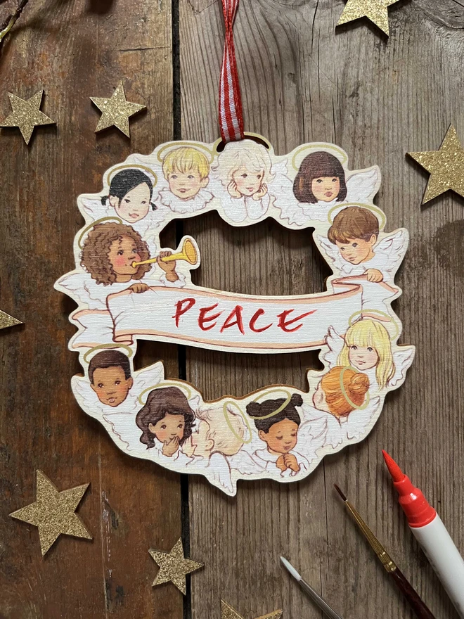 a wooden laser cut printed Christmas decoration featuring a circle of angels drawn in a storybook style with the word PEACE hand written in the angels scroll in the centre