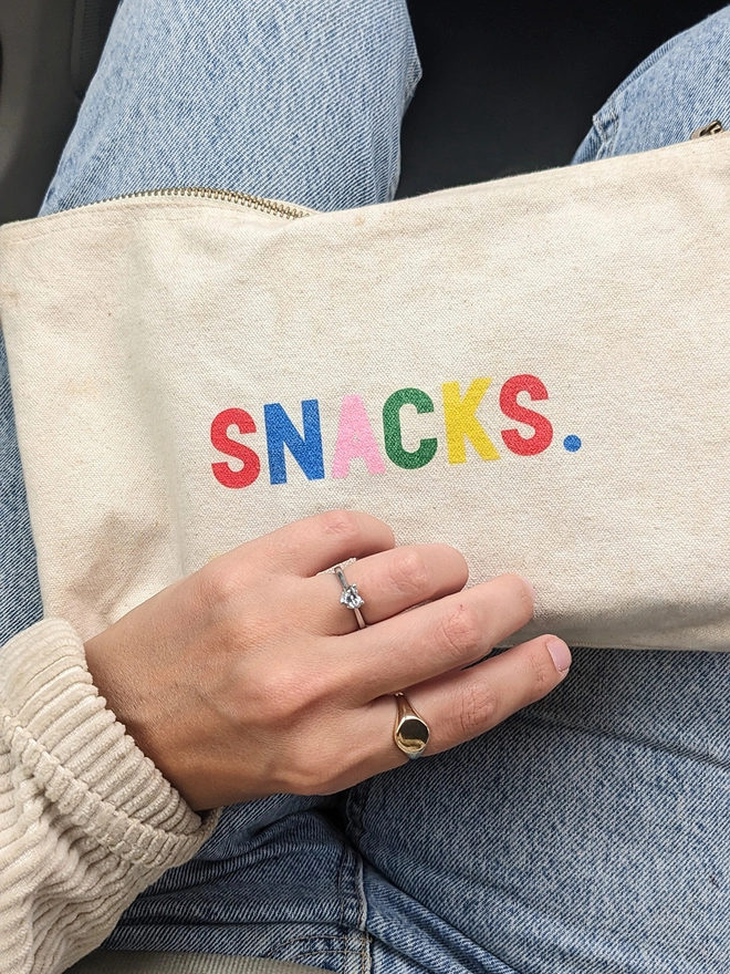 A canvas pouch with multi coloured 'Snacks' slogan on the lap of someone wearing jeans.