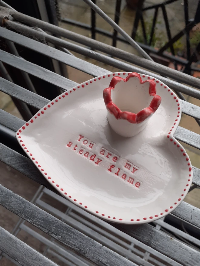 White heart-shaped ceramic candle holder with a red ruffled candle cup and red dotted edge, featuring the message ‘You are my steady flame’, photographed on a metal garden table.