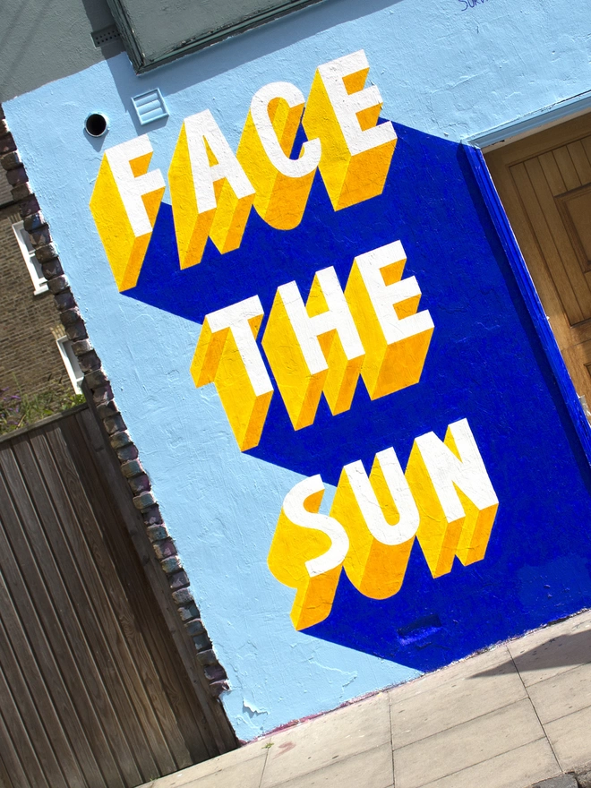 Detail shot of a Survival Techniques street art painting of the words Face The Sun painted in 3d typography in yellow on a blue wall