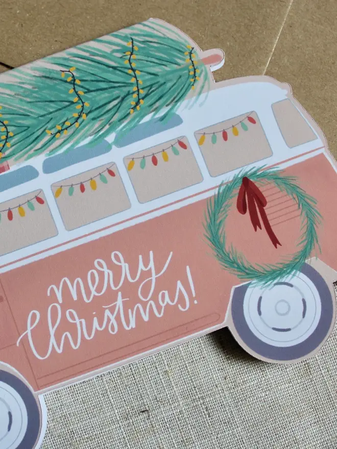 merry christmas illustrated campervan card. campervan shaped card emblazoned with merry christmas and carrying a christmas tree on its roof.