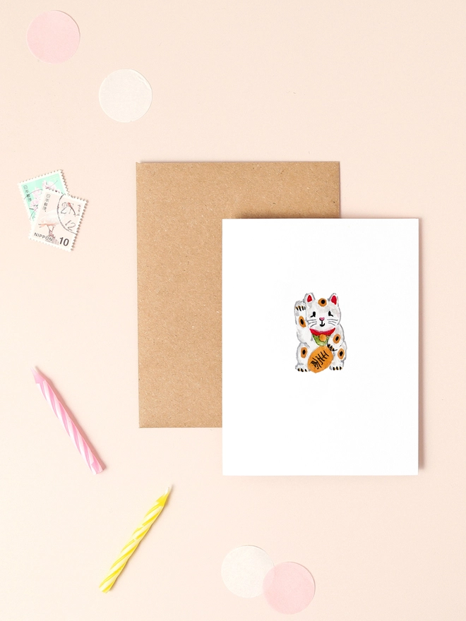 A charming illustration of a traditional Japanese Maneki-neko, or lucky cat, with its paw raised in a welcoming gesture. The cat is adorned in a vibrant red collar with a golden bell, set against a soft, pastel background. The design exudes warmth and positivity, symbolising good fortune and prosperity.