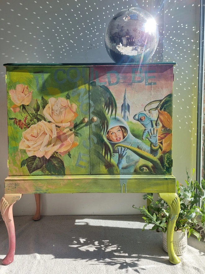 A painted record cabinet decorated with a sci fi image from the 1950s of a lady running from an alien and on the other door, a rose taken from a Victorian seed packet. There is wording that runs down the middle of the cabinet which reads 'it could be worse'. The colours are apple green and pinks.