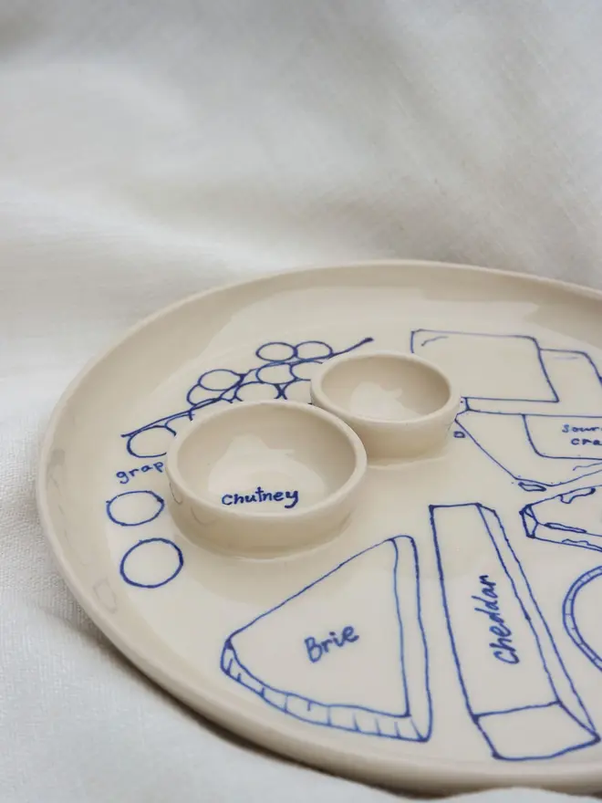 Dippy Cheeseboard Dinner Plate, a white dip plate sitting on a bed cloth. It is covered in blue outlines for cheeses. 