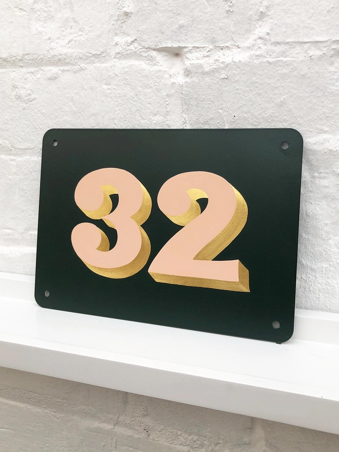 Hand painted house number 32 in pale peach and gold leaf on anthracite grey metal plaque, against a white brick wall. 