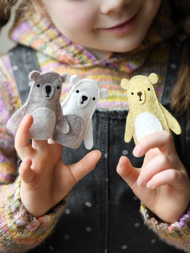 A young child wearing a knitted jumper and grey dungarees holds three handmade felt bear finger puppets on their fingers.