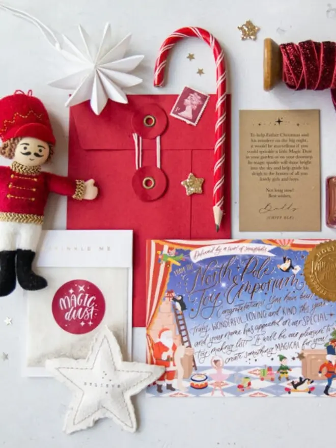 A letter from Father Christmas kit with a red string-and-washer envelope, a kraft Magic Dust instruction card signed by Buddy the chief elf, a "Magic Dust" glitter bag and the illustrated North Pole notecard. Around them are a felt nutcracker, a candy cane pencil, a felt "Believe" star and red glitter ribbon.