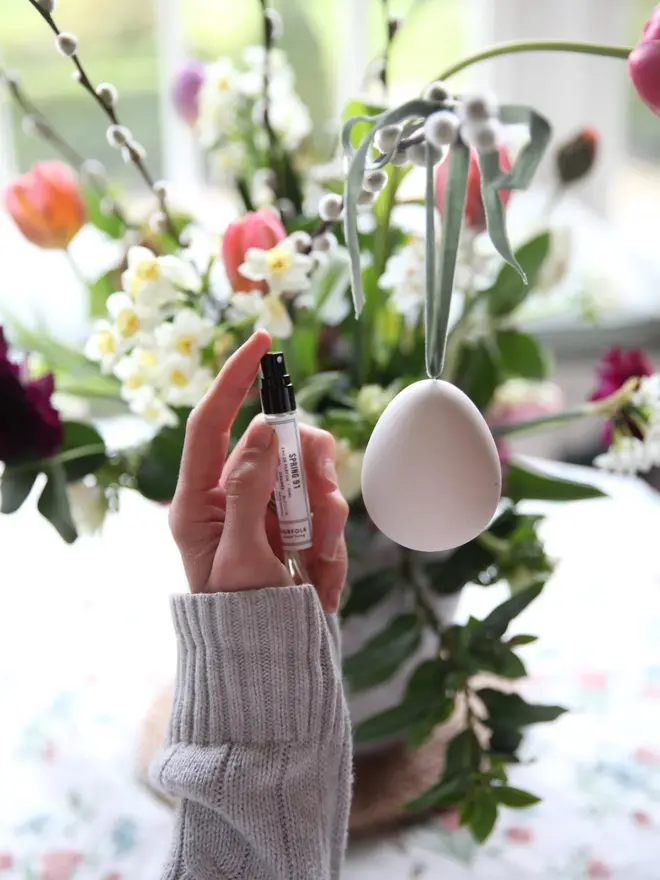 Ceramic egg diffuser with hanging ribbon, being sprayed by a mini perfume vial in a Spring setting