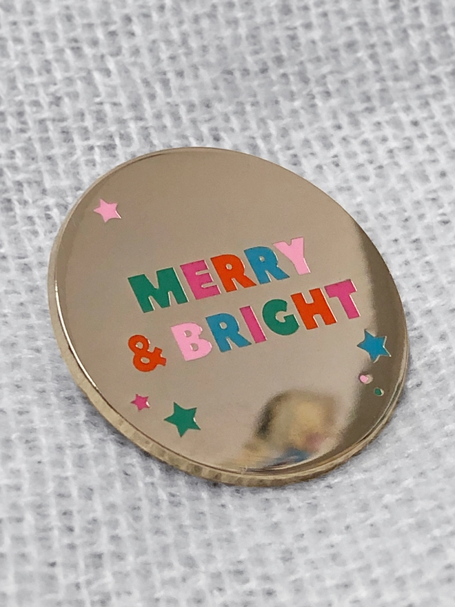 A gold enamel pin with pastel lettering that reads "Merry & Bright" lays on a knitted fabric blanket.