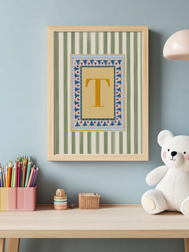 The focal point of this children’s room corner is Annabel Wond Interiors’ signature stripy letter print, featuring a bold yellow “T” on a green and white striped background with a whimsical patterned border. Styled above a cozy wooden desk with playful accents, the print adds a touch of charm and personality to a thoughtfully designed space.