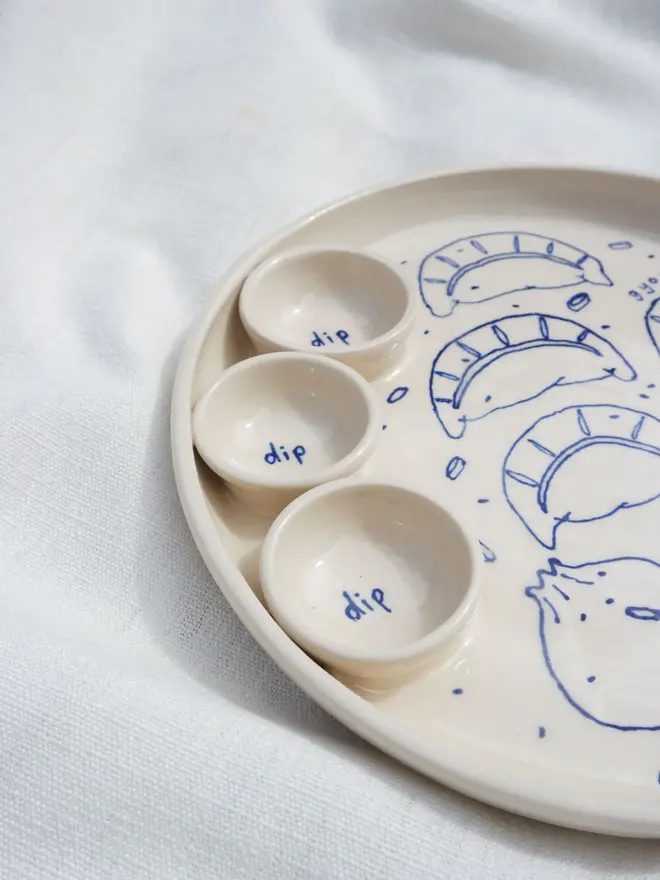 Dumplings Dippy Dinner Plate, a dipping plate sitting on a white bed cloth. It is covered in blue outlines for dumplings. 