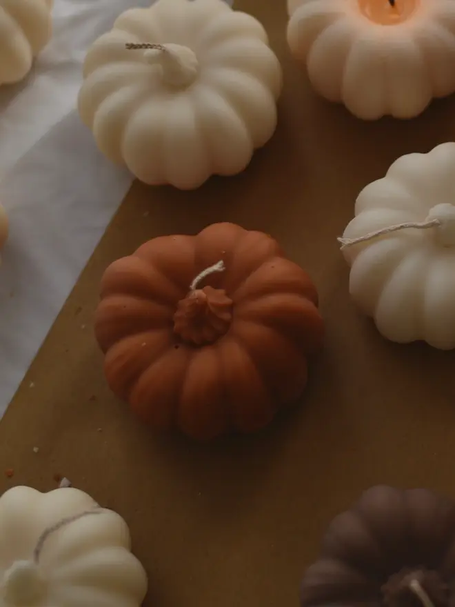 Medium Rust Wax Pumpkin Candle. A top-down view of a terracotta pumpkin candle surrounded by cream and brown pumpkin candles on kraft paper and white linen. The matte sculptural candles add earthy, rustic charm to harvest and Halloween décor.