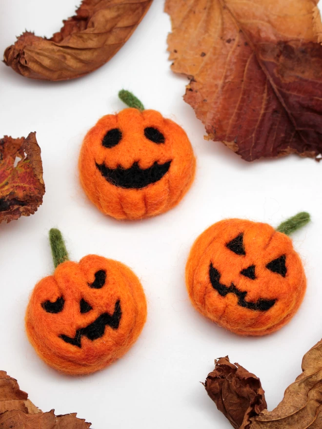 Three needle-felted pumpkins with different expressions. They are on a white background, with autumnal leaves around them