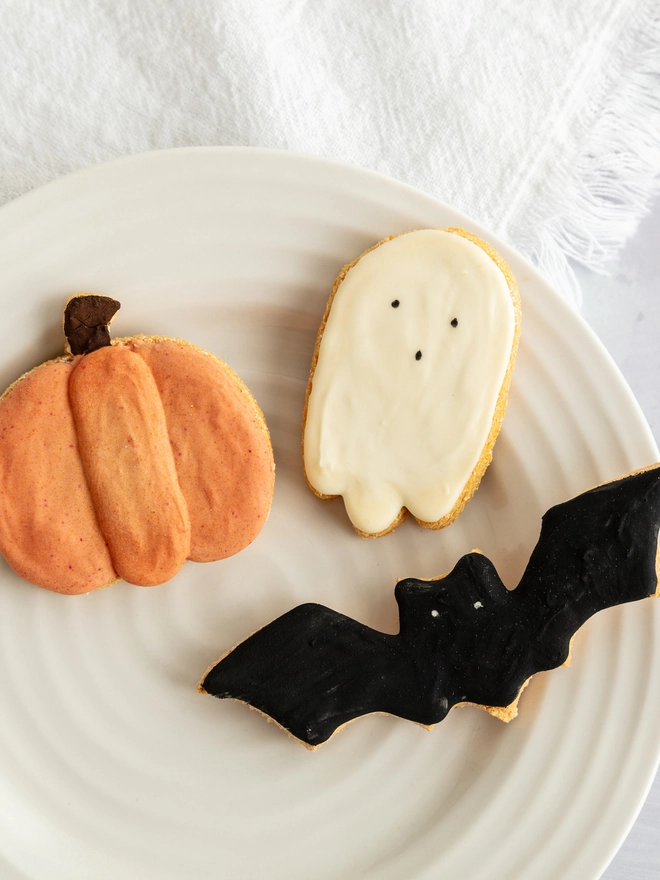 Halloween iced dog biscuit trio featuring a pumpkin, ghost and black bat arranged beside a dog and ceramic bowl.