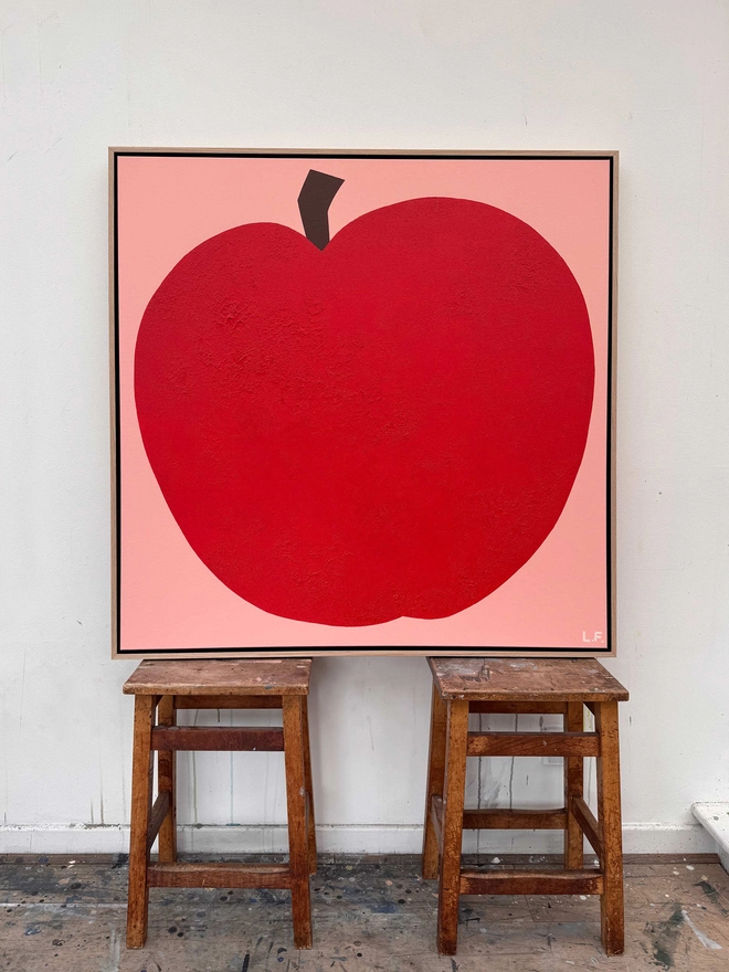 Large painting of a bright red apple on a pink background. Propped on two wooden stools