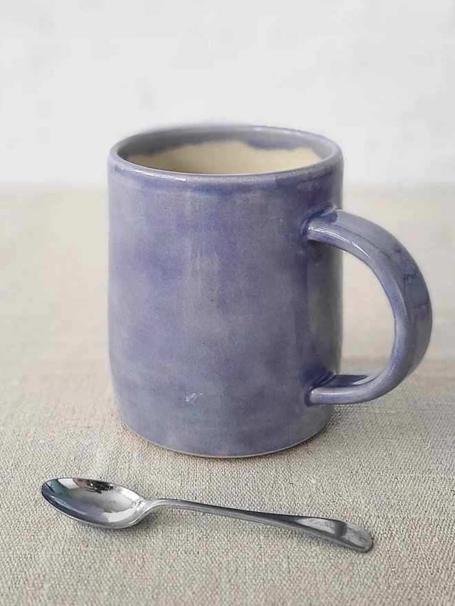 Ceramic Pint Mug, a colourful ceramic mug sitting on a plain surface against a white backdrop. It is accompanied by a spoon. 