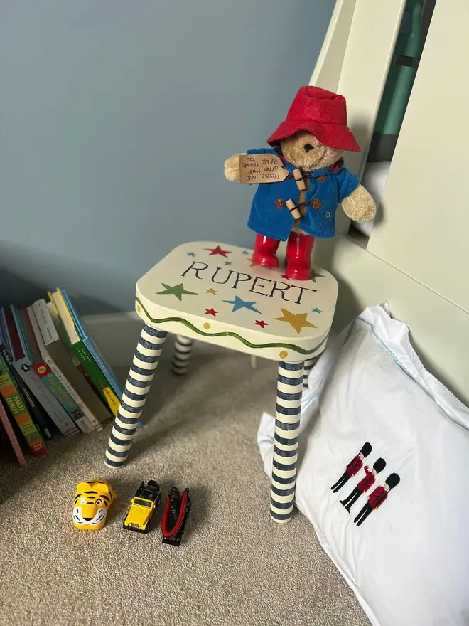 personalised hand-painted nursery stool. a small white stool with blue and white striped legs, painted with 'rupert' on top.