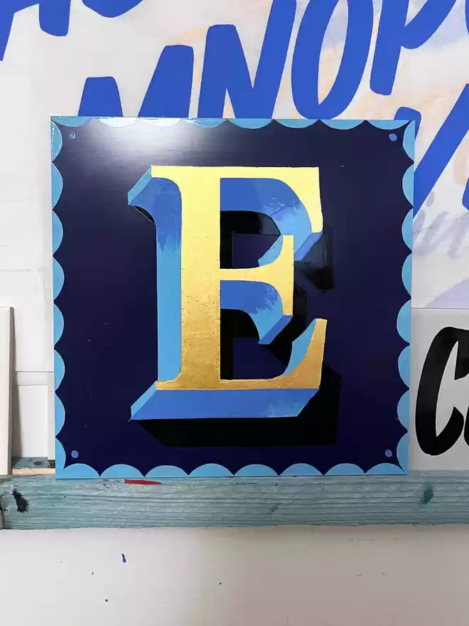 Navy panel with 'E' in thick and thin, wit gold leaf, with light blue close shade and scalloped border.