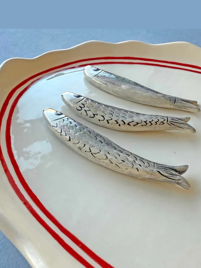 ceramic white platter with red border and anchor detail, with 3 ceramic sardines arranged on top, a ceramic wedge of lemon plus lemon painted on, with parsley detail, on a blue background