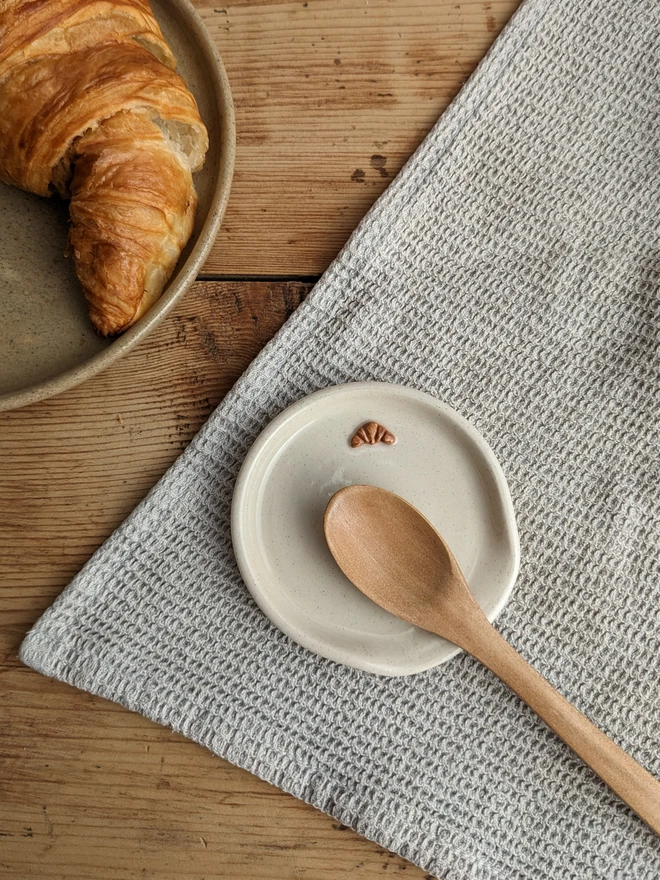 Stoneware spoon rest in vanilla-coloured glaze with a croissant design and a spoon beside it, on a wooden table.