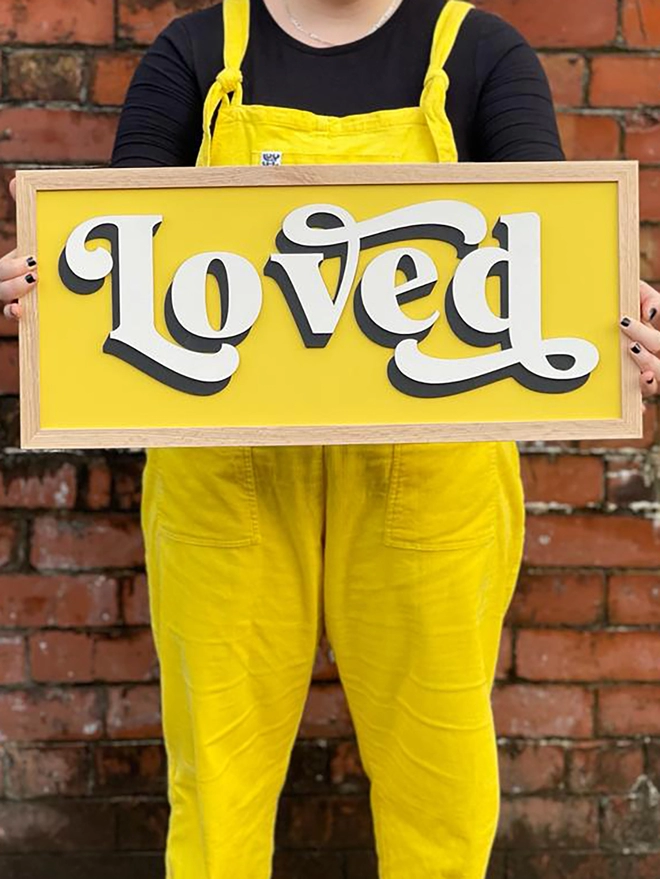 a person wearing yellow dungarees holds a sign which reads LOVED, with a yellow backboard