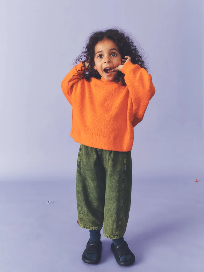 Child pulls a surprised face with hands raised against a lilac backdrop, wearing a Studio Koter orange textured fleece top and green acid-wash barrel-leg jeans.