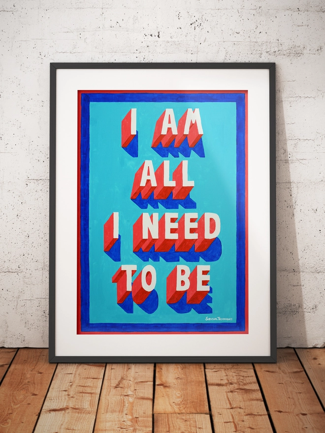 Framed typographic art print standing upright against a white concrete wall on a wooden floor. The design showcases the phrase "I AM ALL I NEED TO BE" in blocky, three-dimensional red and white text with blue shadows, set against a vivid turquoise background and bordered in red and blue. Artist signature "Survival Techniques" appears in the bottom right corner.