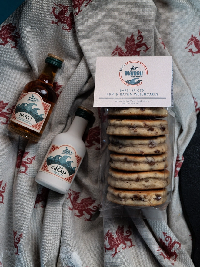 A pack of Barti Rum and Raisin Welsh Cakes, a bottle of miniature Barti Rum and miniature Barti Cream lying on a welsh dragon and beige table cloth