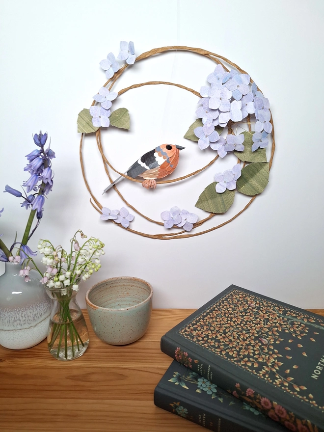 Handmade paper sculpture depicting a chaffinch bird perched on a wreath of baby blue hydrangea flowers and leaves.