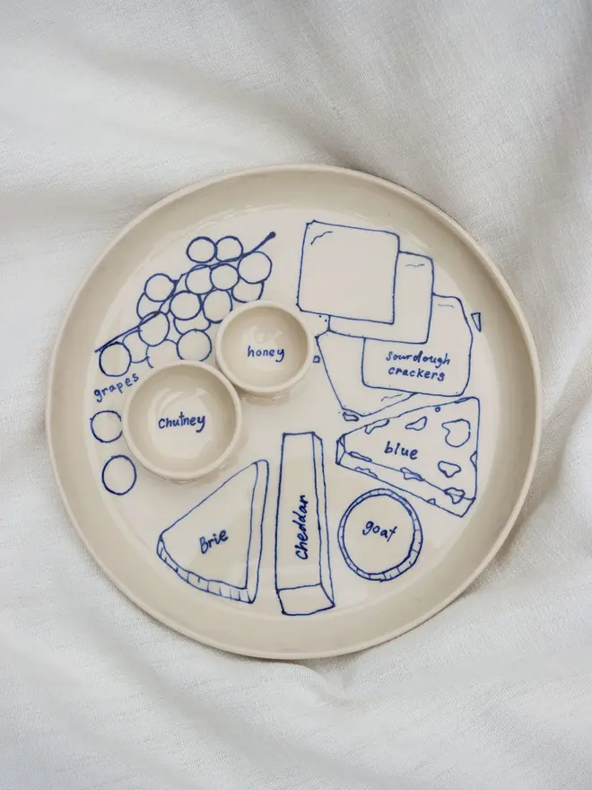 Dippy Cheeseboard Dinner Plate, a white dip plate sitting on a bed cloth. It is covered in blue outlines for cheeses. 