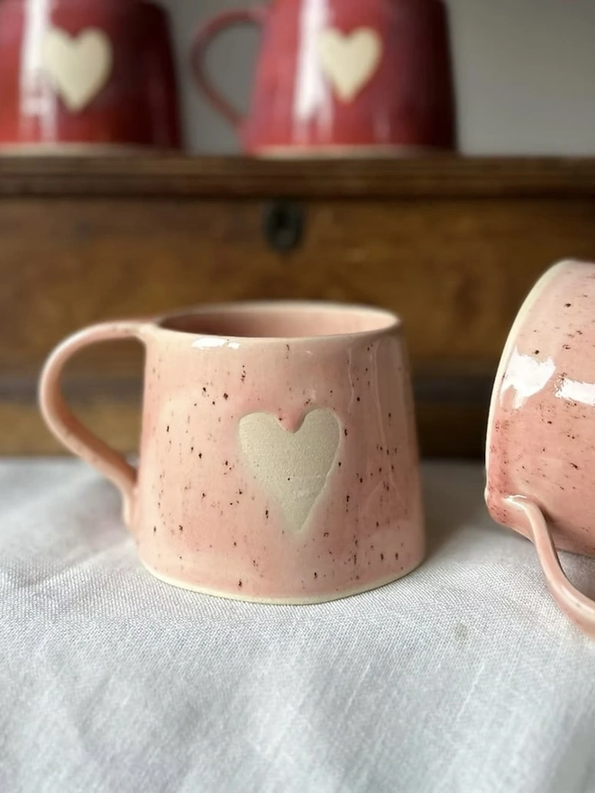 Heart Mug Strawberry Pink, pair of mugs on a white surface in front of two red heart mugs above them. 