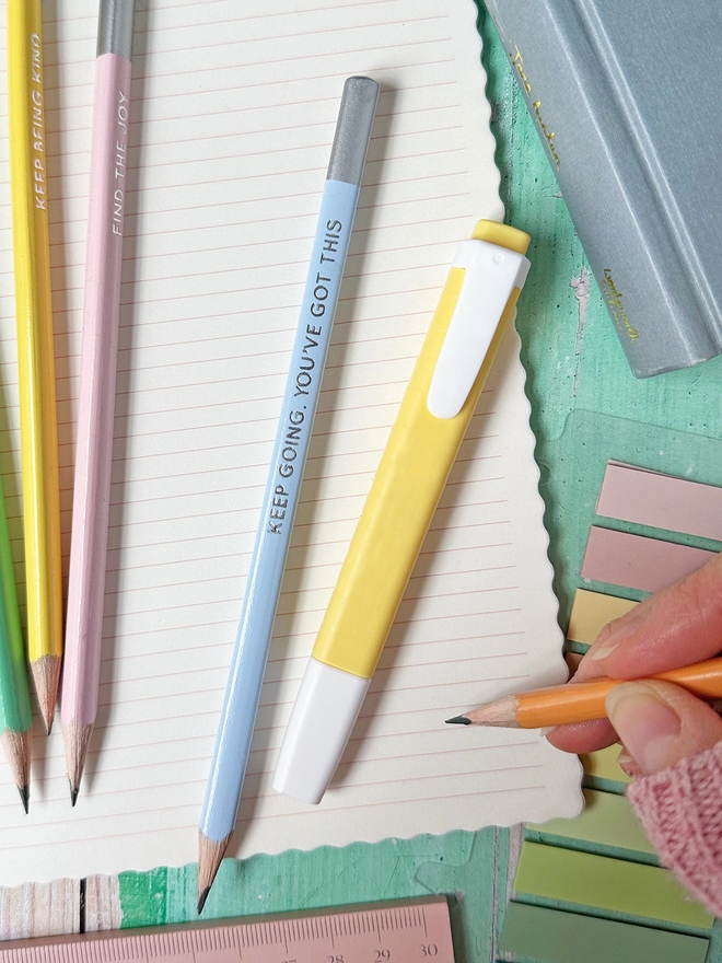 Several pencils and a yellow highlighter lay on an open lined notebook with various other stationery items on a green desk.
