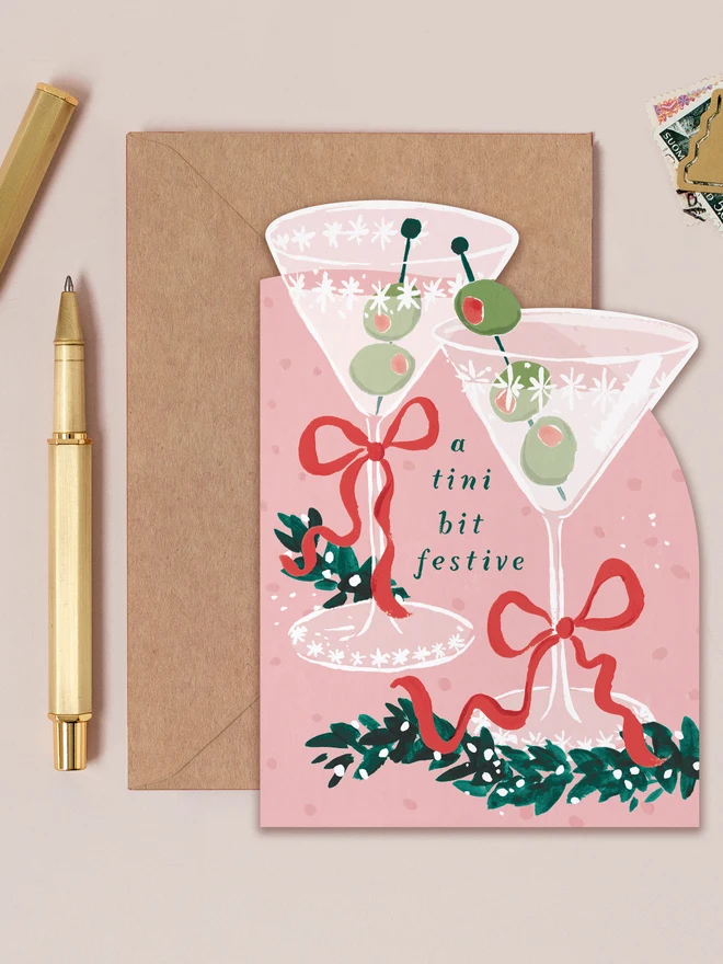 Pink Tini Bit Festive Christmas card shaped like two martini glasses with olives and red bows, lettered 'a tini bit festive', on a kraft envelope beside a gold pen
