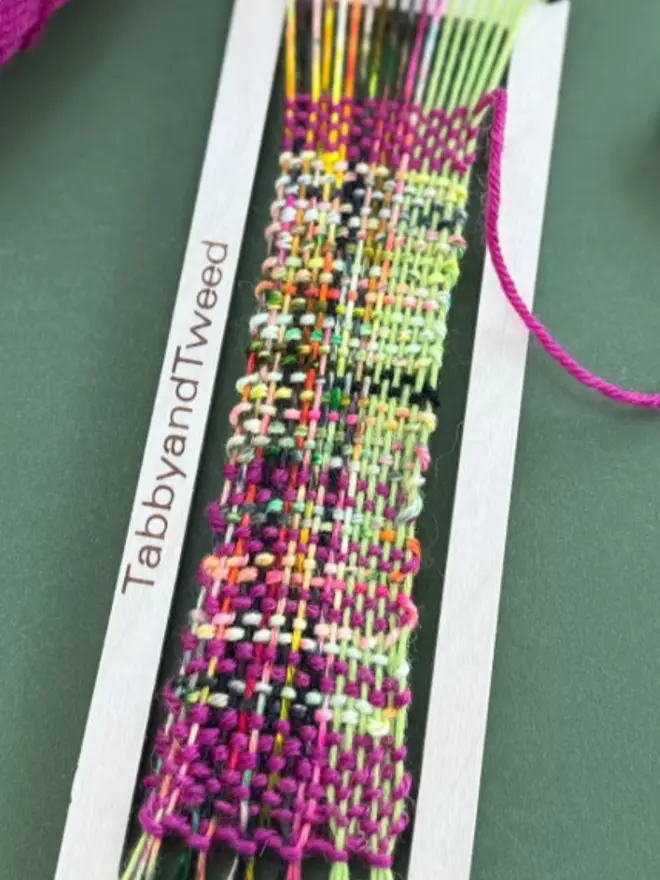 Bright pink and lime bookmark weaving loom kit with multicolored woven sample, yarn skeins, and beige wooden tools on a dark green background.