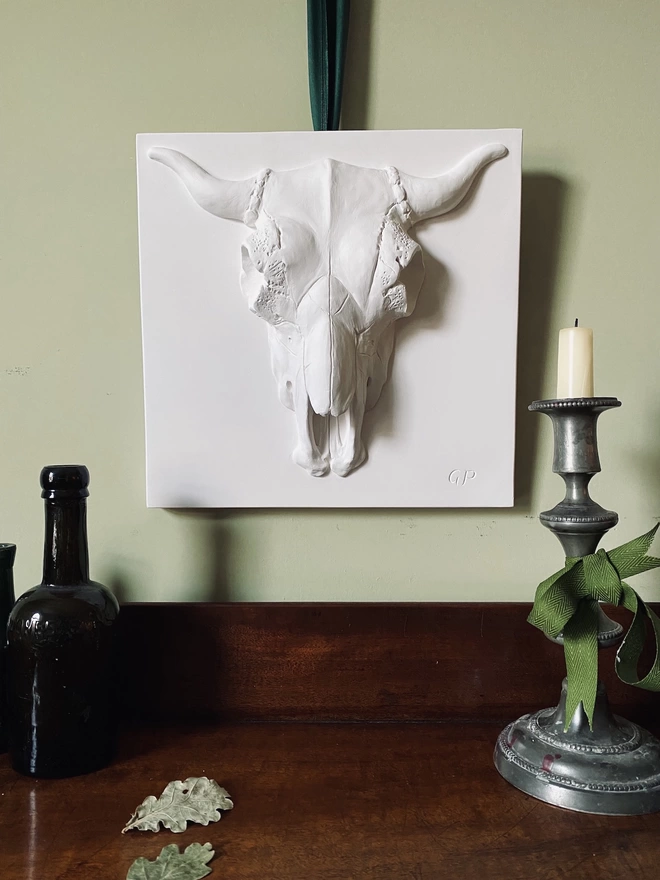Plaster bas-relief wall plaque with Bucranium skull design above a table with bottle and candlestick